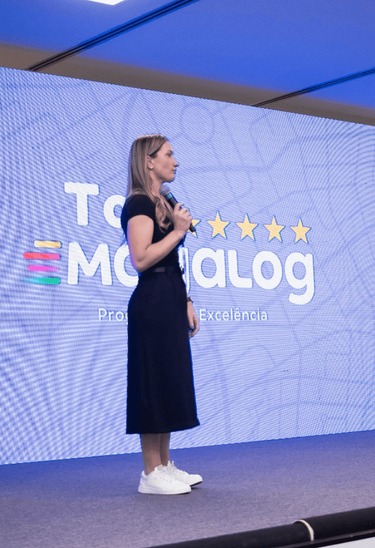 Apresentador no palco do evento Top Magalog, com um grande painel de LED exibindo o logotipo do programa de excelência.