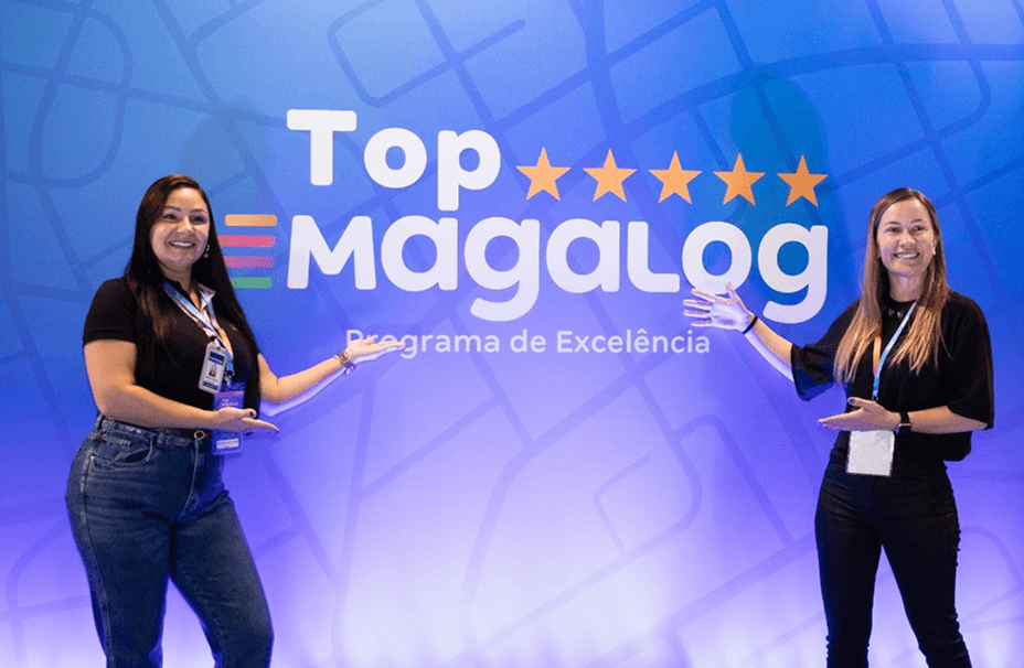 Duas pessoas posando em frente ao painel do evento Top Magalog, com caixas da Magalog empilhadas ao fundo.