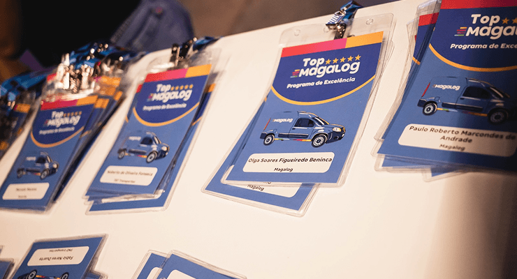 Crachás do evento Top Magalog dispostos sobre uma mesa, com design azul e detalhes do programa de excelência.