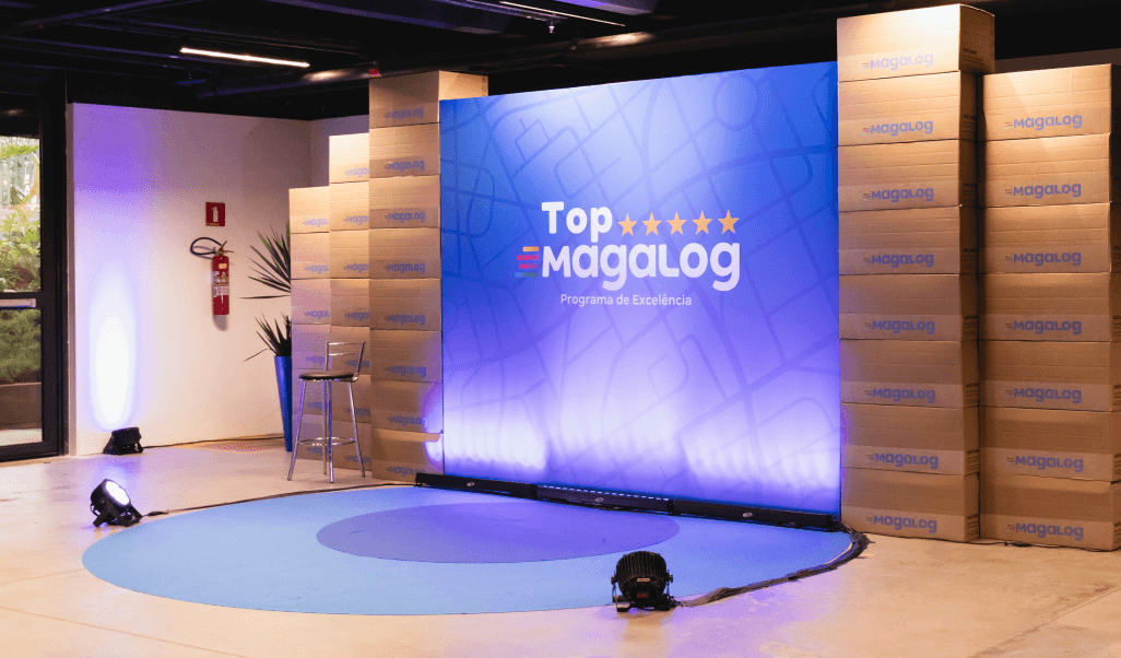 Palco decorado para o evento Top Magalog, com um painel azul exibindo o logotipo do programa e caixas empilhadas com a marca Magalog ao fundo.